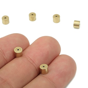 May include: Small, cylindrical, gold-colored metal beads with a hole through the center. Several beads are scattered on a white background, with a few held between fingers. These beads are likely used for jewelry making or other craft projects.