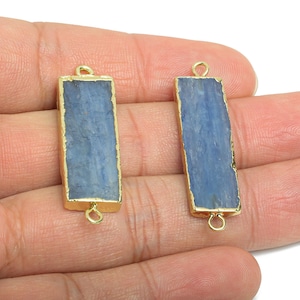 May include: Two rectangular blue kyanite gemstone pendants with gold-coloured edges and loops. The pendants are held in a hand against a white background. The gemstones have a rough, natural texture.