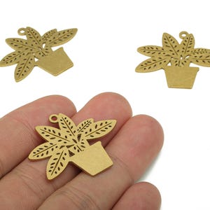 May include: Gold-colored metal charms in the shape of potted plants. The charms feature a detailed cutout design of leaves and a square pot. One charm is held in a hand, with two others on a white background.