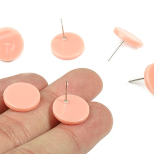 Op de afbeelding: Een verzameling perzikroze, ronde oorstekers met zilveren pinnen. De oorbellen worden op een witte achtergrond getoond, sommige worden in een hand gehouden. De oorbellen zijn gemaakt van een glad, glanzend materiaal.