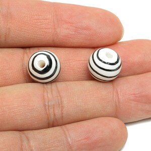 May include: Two round beads with a white base and black spiral stripes. Each bead has a hole through the centre. The beads are held in a hand, and are likely for jewellery making or crafts.