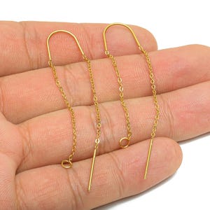 May include: A pair of gold-colored threader earrings. Each earring features a curved top, a delicate chain, and a straight bar at the end. The earrings are displayed against a neutral background.