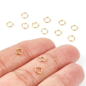 May include: Gold-coloured jump rings, scattered on a white surface and held in a hand. These small, circular rings are used for jewellery making and crafting. The rings are approximately 0.6 cm in diameter.