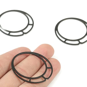 May include: Three black metal jewelry components with a circular design and a geometric accent. One component is held in a hand, while two others are displayed on a white background. These components are suitable for jewelry making.