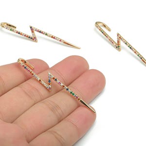 Op de afbeelding: Goudkleurige bliksemvormige oorbellen met een zigzagontwerp. De oorbellen zijn versierd met kleine, kleurrijke edelstenen in een regenboogpatroon. De oorbellen hebben een oorbelhaaksluiting.
