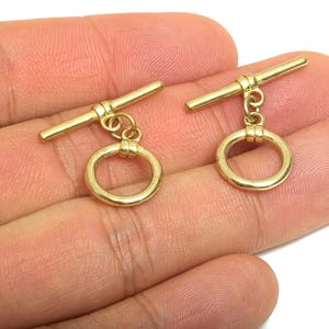 Könnte beinhalten: Zwei goldfarbene Knebelverschlüsse, jeweils mit einem horizontalen Stab und einem kreisförmigen Ring. Die Verschlüsse werden vor einem hautfarbenen Hintergrund gezeigt, der ihr Design und ihre Farbe hervorhebt. Diese Verschlüsse werden wahrscheinlich für die Schmuckherstellung verwendet.