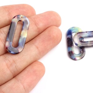 May include: Two oval acrylic beads with a marbled pattern in blue, purple, and yellow hues. One bead is held in a hand, the other is linked to a second bead. Each bead has a small hole for stringing.