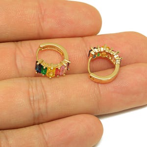 May include: A pair of gold-coloured hoop earrings. Each earring features a row of colourful, rectangular gemstones in a rainbow pattern. The gemstones include blue, green, yellow, pink, and orange hues. The earrings are displayed on a light background.