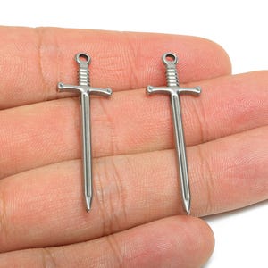 May include: Two small, silver-coloured sword charms. Each charm has a crossguard and a loop at the top. The charms are held in a hand against a white background. These charms are suitable for jewellery making.