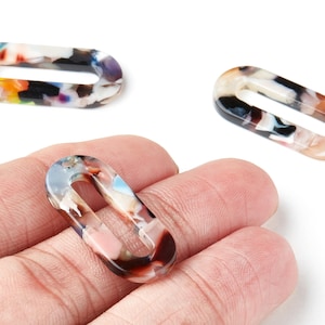 Könnte beinhalten: Drei ovale Kunstharz-Glieder mit offenem Mittelteil in Schildpattmuster. Die Glieder weisen eine Mischung aus Farben auf, darunter Weiß, Schwarz, Braun, Orange und Blau. Ein Glied wird in einer Hand gehalten, während sich zwei weitere auf weißem Hintergrund befinden.