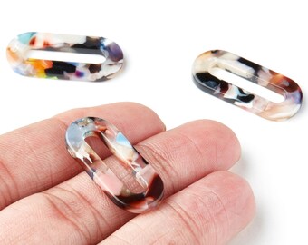 May include: Three oval-shaped, open-center resin links in a tortoiseshell pattern. The links feature a mix of colors including white, black, brown, orange, and blue. One link is held in a hand, while two others are on a white background.