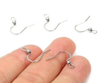 May include: Close-up of several silver-colored earring hooks. The hooks have a curved design with a small ball and a loop at the end. One hook is held by a person's fingers. The hooks are for jewelry making.
