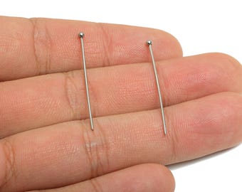 May include: Two silver-colored head pins with a small ball at the top. The pins are held in a hand, and the background is a plain white. These pins are likely used for jewelry making or other craft projects.