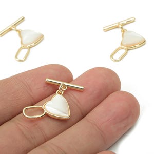 May include: Gold-colored metal toggle clasp with a heart-shaped, pearlescent charm. The clasp features a bar and an oval loop, perfect for jewelry making. The heart charm has a white, iridescent finish.
