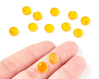 May include: A collection of small, round, amber-colored beads with a hole in the center. The beads are scattered on a white surface, with a few resting in a person's hand. These beads are likely used for jewelry making or crafting.