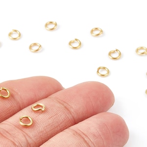 May include: Close-up of small, gold-coloured jump rings. Several rings are scattered on a white surface, with a few resting on a person's fingers. These jewellery findings are used for connecting jewellery components.