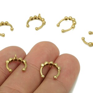 Könnte beinhalten: Goldfarbene Ohrklemmen mit Perlendesign. Das Bild zeigt mehrere dieser Ohrklemmen, einige mit einer kleinen Schlaufe oben. Die Klemmen befinden sich in verschiedenen Positionen, einige werden von einer Hand gehalten.
