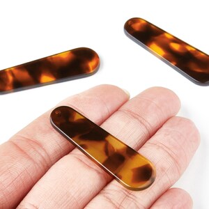 May include: Three oblong, tortoiseshell-patterned beads with rounded ends and a small hole at one end. One bead is held in a hand, while the other two are scattered on a white background. The beads have a glossy finish.