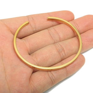 Puede incluir: Una pulsera de brazalete abierta de color dorado. La pulsera tiene un diseño circular simple con un acabado liso y pulido. La pulsera se sostiene en una mano sobre un fondo blanco.