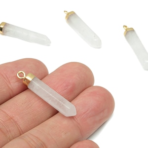 May include: Four clear quartz crystal pendants with gold-coloured caps and loops. One pendant is held in a hand. The crystals are cylindrical with a pointed end. The pendants are approximately 3.8 cm long.