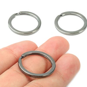 May include: Three gunmetal-colored key rings. One key ring is held in a hand. The key rings are circular with a split for attaching keys. The key rings are made of metal and have a smooth finish. The background is white.