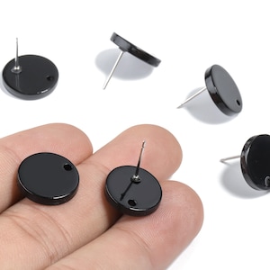 Pode incluir: Brincos de botão redondos pretos com pinos prateados. Os brincos têm um acabamento brilhante e um pequeno orifício perto do pino. Vários brincos estão espalhados sobre um fundo branco.
