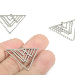 Pode incluir: Três pingentes triangulares geométricos prateados com um design aberto. Os pingentes têm um pequeno laço na parte superior para prender a joias. Um pingente é segurado em uma mão.