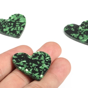 Op de afbeelding: Drie hartvormige decoratieve versieringen met een zwarte basis en een groen, gefragmenteerd patroon. Eén hart wordt in een hand gehouden. De harten zijn waarschijnlijk voor knutselen of het maken van sieraden.