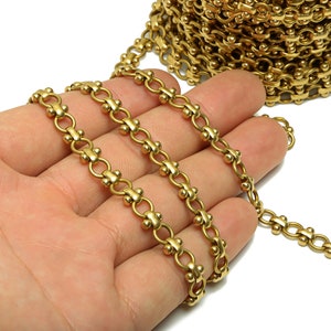 Op de afbeelding: Close-up van een goudkleurige ketting met een herhalend patroon van kleine kralen en gebogen schakels. De ketting is gedeeltelijk opgerold, met een deel ervan in een hand. De ketting wordt waarschijnlijk gebruikt voor sieraden of knutselen.
