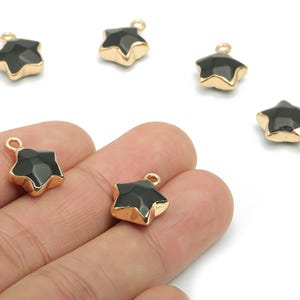 Op de afbeelding: Zwarte stervormige bedels met goudkleurige randen en lussen voor het maken van sieraden. Verschillende bedels zijn verspreid op een witte achtergrond, met twee in een hand. De bedels hebben een facetgeslepen oppervlak.