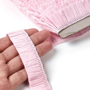 May include: Close-up of a roll of pale pink fringe trim. The fringe is made of a crinkled paper-like material and is held in a hand. The trim is on a white background.