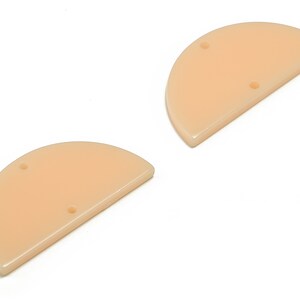 Może przedstawiać: Dwa półkoliste kształty akrylowe w kolorze brzoskwiniowym z dwoma otworami każdy. Kształty są płaskie i gładkie.