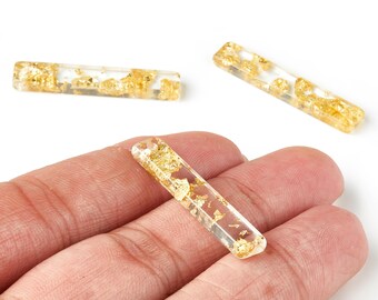 Puede incluir: Tres cuentas rectangulares transparentes de resina con inclusiones de pan de oro. Una de las cuentas se sostiene en una mano. Las cuentas tienen bordes redondeados y el pan de oro añade un toque de lujo.