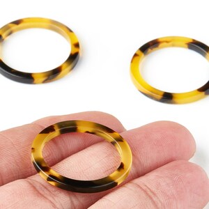May include: Three oval-shaped resin rings with a tortoiseshell pattern of yellow, brown, and black. The rings are shown against a white background, with one held in a hand.