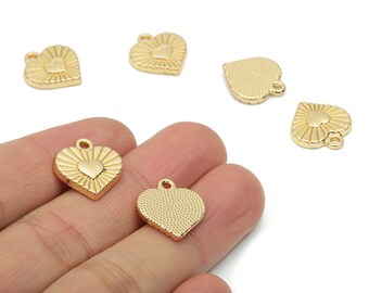 May include: Gold-colored heart-shaped charms with a textured design. The charms feature a smaller heart in the center and a loop for attaching to jewelry. Several charms are visible, some held in a hand.