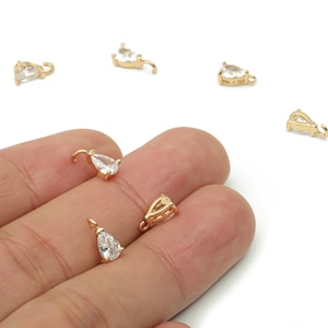 Puede incluir: Pequeños colgantes dorados en forma de lágrima con piedras de circonita cúbica transparente. Los colgantes tienen un lazo en la parte superior para sujetarlos a las joyas. Varios están dispersos sobre un fondo blanco.