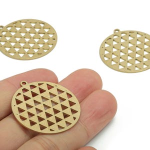 Pode incluir: Três pingentes circulares dourados com um padrão geométrico de triângulos. Um pingente é segurado em uma mão. Cada pingente tem um pequeno laço na parte superior para prender a uma corrente ou outro componente de joia.