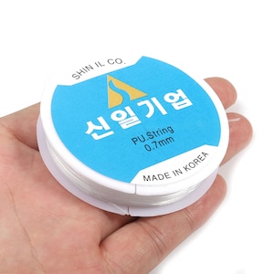 May include: A spool of clear PU string, 0.7mm thick, held in a hand. The spool has a light blue label with Korean text and a gold logo. The text "MADE IN KOREA" is also visible.