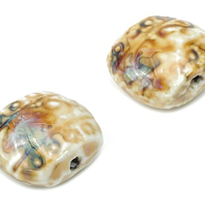 Pode incluir: Duas contas de cerâmica marrom e branca com um furo no centro. As contas têm um padrão manchado e marmorizado.