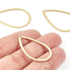 Puede incluir: Tres hallazgos de joyería dorados en forma de lágrima. Uno se sostiene en una mano, mostrando su tamaño y forma. Los hallazgos tienen un pequeño agujero en la parte superior para sujetarlos. Ideales para la fabricación de joyas.