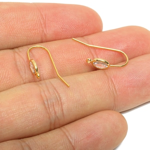 May include: A pair of gold-coloured earrings, each featuring a clear, oval-cut stone. The earrings are presented on a hand, highlighting their intricate design and the sparkle of the stones. The hooks are curved.