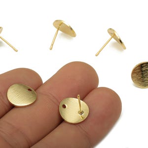 May include: Gold-colored, round earring posts with a textured surface. The earrings have a post and a small hole for attachment. The image shows several earrings, some with the post attached and some without.