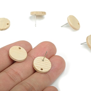 May include: Several unfinished, round wooden stud earrings with silver posts are displayed. The earrings are light beige with a visible wood grain. Each earring has a small hole near the edge. The earrings are shown on a white background.