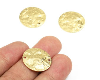 Könnte beinhalten: Drei goldfarbene Metall-Scheibenanhänger mit gehämmertem Finish. Einer wird in einer Hand gehalten, zwei liegen auf weißem Hintergrund. Jede Scheibe hat eine strukturierte Oberfläche und zwei kleine Löcher.