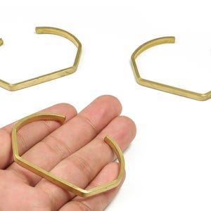 May include: Three gold-colored geometric cuff bracelets. One bracelet is being held in a hand. The bracelets have a flat bar in the center and angled sides, creating a modern, minimalist design. The bracelets are open at the ends.