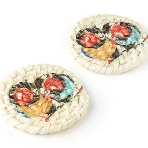 Puede incluir: Dos botones redondos, tejidos, blancos con un patrón floral colorido. Los botones tienen un diseño tejido y están decorados con un patrón floral en tonos rojos, azules y amarillos.