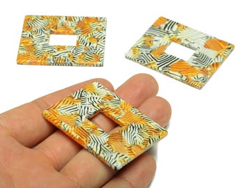 May include: Three decorative rectangular beads with a central square cutout. The beads feature a colorful pattern of orange, black, and white stripes and shapes. One bead is held in a hand, showcasing the size and design.