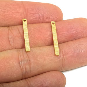 May include: Two gold-colored rectangular metal bars with a textured surface and a small hole at the top. The bars are held in a hand against a white background. These are likely jewelry making components.
