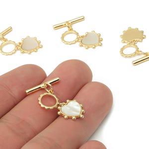 Peut inclure: Fermoirs à bascule dorés avec un charme en forme de cœur et un charme rond. Le charme en forme de cœur a un centre blanc nacré et est entouré de petites perles dorées. Les fermoirs sont présentés sur fond blanc.