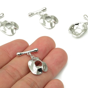 May include: Silver-toned boxing glove charms with a T-bar clasp. The charms are displayed on a white background, with one held in a hand. The charms have a shiny, reflective surface and are likely made of metal.